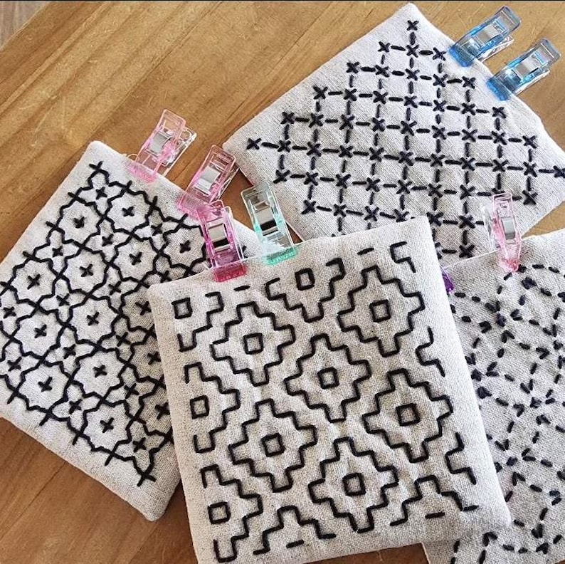 May include: Four square pieces of light beige fabric featuring black hand-stitched embroidery patterns. Each fabric square is secured with colorful plastic sewing clips in shades of pink, blue, and green, displaying geometric designs.