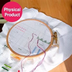May include: A wooden embroidery hoop holding white fabric with various colored stitched patterns. The fabric has a grid pattern with Korean text and the word "Embroidery". Purple thread is threaded through the needle.