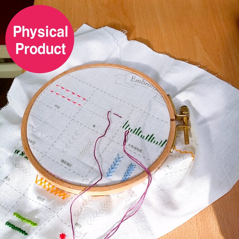 May include: A wooden embroidery hoop holding white fabric with various colored stitched patterns. The fabric has a grid pattern with Korean text and the word "Embroidery". Purple thread is threaded through the needle.