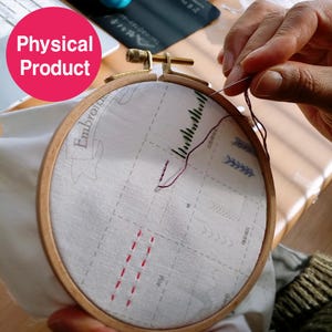 May include: A wooden embroidery hoop holding white fabric with a printed design. Red and black thread is being used to stitch the fabric. The text "Embroider" is visible on the fabric. The image is labeled "Physical Product".