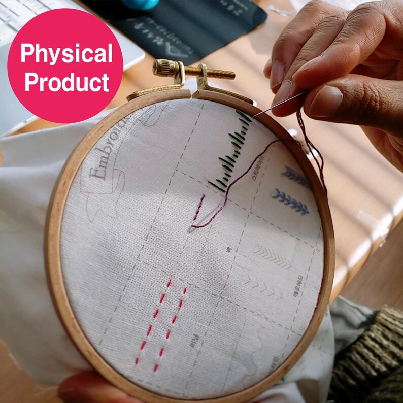 May include: A wooden embroidery hoop holding white fabric with a printed design. Red and black thread is being used to stitch the fabric. The text "Embroider" is visible on the fabric. The image is labeled "Physical Product".