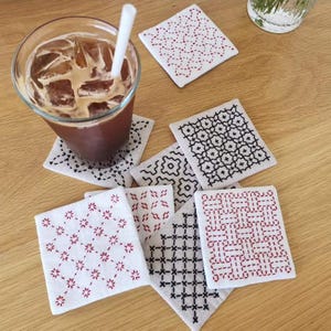 May include: A collection of square fabric coasters featuring intricate hand-stitched embroidery patterns in red and black thread on white fabric, alongside a clear glass filled with iced coffee and a white straw, arranged on a wooden surface.