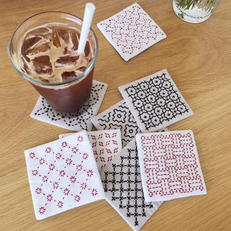 May include: A collection of square fabric coasters featuring intricate hand-stitched embroidery patterns in red and black thread on white fabric, alongside a clear glass filled with iced coffee and a white straw, arranged on a wooden surface.