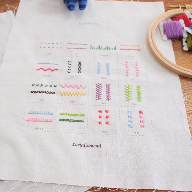 May include: A white fabric with various embroidery stitch examples in different colors. The fabric has the text "Embroidery" at the top and "Lucydiamond" at the bottom. A wooden embroidery hoop and spools of thread are in the background.