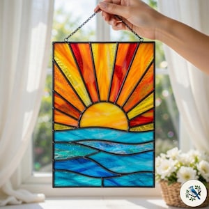 Può includere: Pannello in vetro colorato raffigurante un tramonto sull'oceano. Il sole è giallo brillante, con raggi arancioni e rossi. L'oceano è blu e turchese. Il pannello è incorniciato in nero e tenuto da una catena.