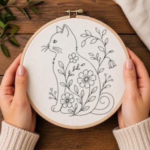 May include: A circular wooden embroidery hoop containing white fabric with a black line art design of a cat sitting in profile. The cat's body is filled with floral and botanical patterns, including various flowers, leaves, and stems.