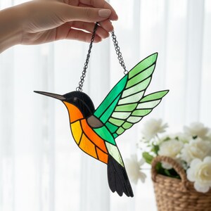 May include: A stained glass hummingbird suncatcher, suspended by a black chain. The bird has a black head, an orange and yellow body, and green wings. The wings are a gradient of light and dark green. The hummingbird's beak is long and dark.