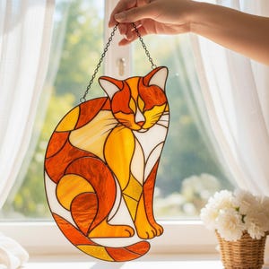 Può includere: Decorazione a forma di gatto in vetro colorato nei toni dell'arancione, giallo e bianco. Il gatto è seduto, con una catena per appenderlo. L'arte della finestra è esposta davanti a una finestra con tende bianche e un cesto di fiori bianchi.