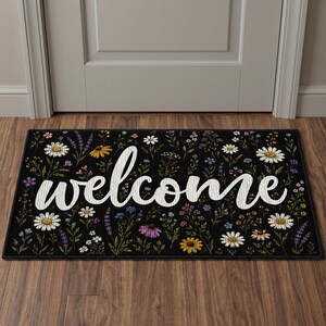 May include: A rectangular black doormat featuring a colorful botanical print of various wildflowers including white daisies, yellow blossoms, and purple sprigs. The word welcome is printed across the center in a large, white, cursive script font.