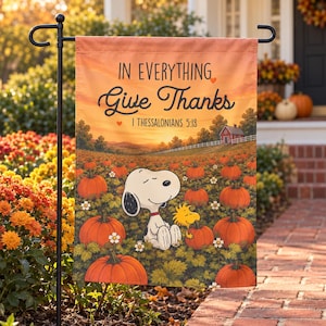May include: A rectangular garden flag featuring a sunset pumpkin patch scene with Snoopy and Woodstock. The text reads IN EVERYTHING Give Thanks 1 THESSALONIANS 5:18. The design includes orange pumpkins, green foliage, and a red barn.