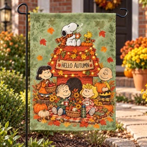 May include: A rectangular garden flag featuring a green background with falling autumn leaves. Snoopy and Woodstock sit on a red doghouse roof. Below, Lucy, Linus, Sally, and Charlie Brown gather with pumpkins, a basket, and mugs. Text reads HELLO AUTUMN.