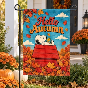 May include: A rectangular garden flag featuring the text Hello Autumn in orange and red letters. The illustration shows a white dog and a yellow bird resting on a red doghouse, surrounded by falling orange and red maple leaves and acorns.