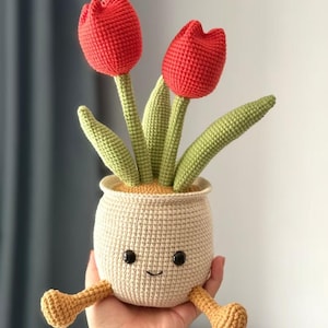 Könnte beinhalten: Gehäkelte Tulpenpflanze mit zwei roten Blüten, grünen Blättern und einem beigefarbenen Topf mit einem lächelnden Gesicht und kleinen Beinen. Die handgefertigte Pflanze wird in einer Hand gehalten.