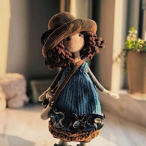 Könnte beinhalten: Handgefertigte Häkelpuppe mit braunen Haaren, braunem Hut und blauem Kleid. Die Puppe hat eine kleine Tasche und Schuhe. Die Puppe steht in der Nähe eines Fensters.