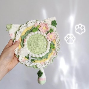 Könnte beinhalten: Ein handgefertigter, gehäkelter Untersetzer in Katzenform in Grün-, Weiß-, Gelb- und Rosatönen. Der Untersetzer hat einen Mittelkreis, florale Akzente und eine baumelnde Glocke. Die Ohren sind dreieckig und das Design ist detailliert.