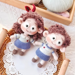 Könnte beinhalten: Zwei handgefertigte Häkelpuppen mit lockigem braunem Haar, rosigen Wangen und großen Augen. Sie tragen blau-weiß gestreifte Oberteile und blaue Röcke. Eine Puppe hat rote Schleifen. Die Puppen befinden sich auf einer weißen Spitze in einem Weidenkorb.