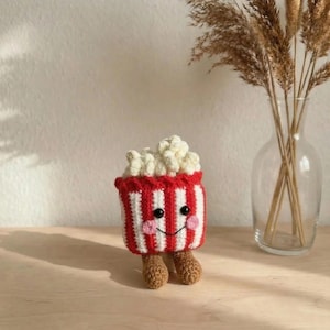 Könnte beinhalten: Ein gehäkelter Popcornbehälter mit einem lächelnden Gesicht, schwarzen Augen und rosa Wangen. Der Behälter ist rot mit weißen Streifen und mit weißem Popcorn gefüllt. Er hat braune gehäkelte Füße.