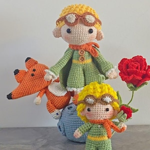 Könnte beinhalten: Handgefertigte Häkelpuppen des kleinen Prinzen, eines Fuchses und einer roten Rose. Die Puppen des kleinen Prinzen tragen grüne Outfits, orangefarbene Schals und eine Brille. Der Fuchs ist orange mit schwarzen Akzenten.