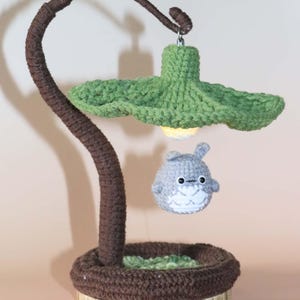 May include: A handmade crochet lamp featuring a brown stem and base, a green leaf-shaped lampshade, and a gray Totoro character. The lamp has a warm, inviting glow, perfect for a child's room or a whimsical decor theme.