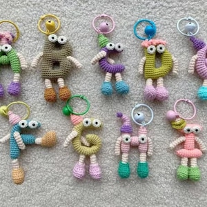 Könnte beinhalten: Eine Sammlung handgefertigter Häkel-Schlüsselanhänger in Form von Buchstaben und Zahlen. Jeder Schlüsselanhänger hat Wackelaugen, Arme, Beine und eine kleine Glocke oder einen Anhänger, der an einem Ring befestigt ist. Die Farbpalette umfasst Grün, Braun, Lila, Rosa, Blau und Gelb.