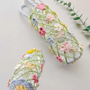 Könnte beinhalten: Zwei klare Glasflaschen sind in gehäkelten Flaschenhaltern eingeschlossen. Die Halter sind hellgrün mit einem Blumenmuster aus rosa, blauen, gelben und weißen Blüten. Ein Halter hat einen Griff, der andere ist eine kleinere Version.