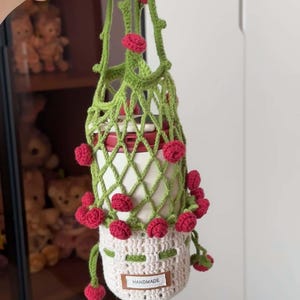 Könnte beinhalten: Ein handgefertigter Wasserflaschenhalter aus Häkelarbeit in Weiß und Grün, verziert mit roten Blumenakzenten. Der Halter hat ein Gittermuster und ein kleines Etikett mit der Aufschrift "HANDMADE". Die Flasche im Inneren ist weiß mit einem roten Deckel.