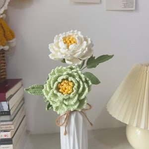 Könnte beinhalten: Eine weiße Keramikvase hält zwei gehäkelte Blumen. Eine ist weiß mit gelbem Zentrum, die andere hellgrün mit gelbem Zentrum. Grüne Blätter sind befestigt. Die Vase ist mit Bindfaden gebunden.