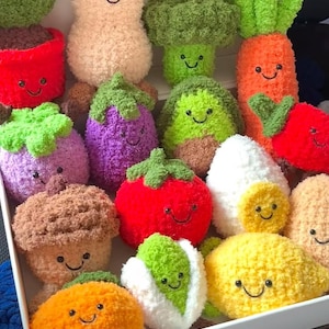 Könnte beinhalten: Eine Sammlung von Stofftieren in Form von lächelndem Gemüse, darunter eine Karotte, Brokkoli und Tomate. Die Spielzeuge bestehen aus weichem, strukturiertem Stoff in leuchtenden Farben. Sie sind in einer weißen Box angeordnet.