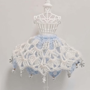 Könnte beinhalten: Eine Miniatur-Kleiderpuppe in Weiß zeigt einen handgefertigten Häkelrock. Der Rock hat ein hellblaues und weißes Design mit Schneeflocken- und Perlenverzierungen. Die Häkelarbeit hat ein zartes, offenes Muster.