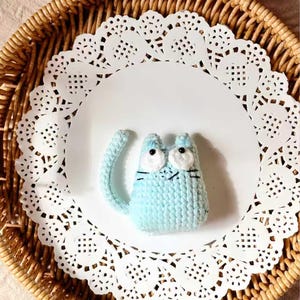 May include: A light blue crocheted cat figurine with white eyes and black whiskers sits on a white paper doily. The doily is placed inside a woven wicker basket. The cat has a long tail.