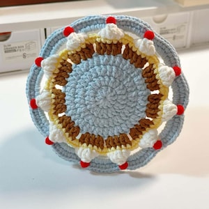 Könnte beinhalten: Ein handgefertigter, gehäkelter Untersetzer in einem kreisförmigen Kuchen-Design. Der Untersetzer hat eine hellblaue Mitte, einen Ring aus Braun und Gelb sowie weiße cupcake-ähnliche Verzierungen mit roten Kirschakzenten am Rand. Der Untersetzer schützt Oberflächen vor heißen oder kalten Getränken.