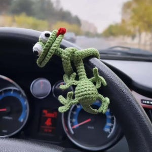 Op de afbeelding: Een groen gehaakt kikker auto-accessoire met witte ogen en een rode tong is bevestigd aan een zwart stuurwiel. De kikker is gemaakt van garen en heeft een grillig ontwerp. De achtergrond toont een wazig zicht op een weg.