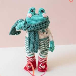 Könnte beinhalten: Eine handgefertigte Häkelfroschpuppe mit türkisfarbenem Kopf und Schal, die einen cremefarbenen Pullover und gestreifte Hosen trägt. Der Frosch hat rote Stiefel und rosa Wangen. Die Puppe ist ein skurriles, handgefertigtes Produkt.