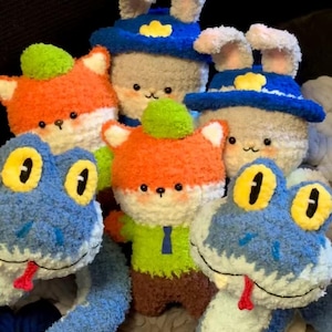 Könnte beinhalten: Eine Sammlung handgefertigter Plüschtiere mit verschiedenen Tierdesigns. Die Spielzeuge umfassen Füchse in Orange und Weiß, Kaninchen mit blauen Hüten und blaue Schlangen. Jedes Spielzeug hat gestickte Details und ein weiches, strukturiertes Aussehen.
