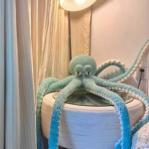 Könnte beinhalten: Ein großer, handgefertigter Häkel-Oktopus in Türkis- und Weißtönen. Der Oktopus sitzt auf einem runden, weißen Tisch mit Glasplatte. Die Tentakel hängen herunter, und die Augen sind groß und ausdrucksstark. Über dem Oktopus befindet sich eine Lampe mit weißem Schirm.