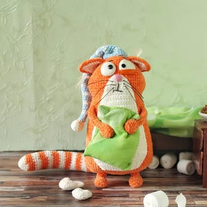 Op de afbeelding: Een handgemaakte oranje en witte gehaakte kattenpop met een blauw-witte nachtmuts en een groen kussen. De kat heeft grote, wijde ogen en een gestreepte staart. De pop is een grillig, zacht speelgoed.