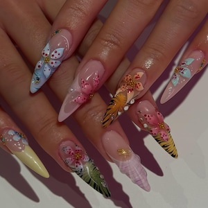 Puede incluir: Uñas largas y puntiagudas con intrincados diseños florales. Cada uña presenta un diseño diferente, incluyendo flores, rayas de tigre y otros elementos decorativos. Las uñas están pintadas en varios colores, incluyendo rosa, amarillo, azul y naranja, con detalles dorados.