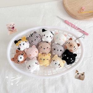 Może przedstawiać: Biały kosz wypełniony różnymi szydełkowymi ozdobami w kształcie kota. Koty są w różnych kolorach, w tym białym, czarnym, brązowym, żółtym i różowym, z haftowanymi rysami twarzy. Szydełko i drewniana taca znajdują się w tle.