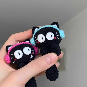 Pode incluir: Duas figuras de gato de crochê preto feitas à mão com olhos brancos grandes, protetores de ouvido rosa e azul pequenos e pernas pretas pequenas. As figuras de gato são seguradas na mão de uma pessoa contra um fundo neutro. Os gatos têm pequenos bigodes brancos e bocas vermelhas.