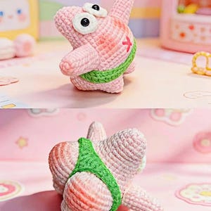 Könnte beinhalten: Handgefertigtes, rosa gehäkeltes Seestern-Spielzeug mit weißen Augen und einem grünen Band. Der Seestern hat ein rotes 'X' auf seinem Körper. Das Spielzeug wird auf einer rosa Oberfläche mit anderen farbenfrohen Objekten im Hintergrund präsentiert.