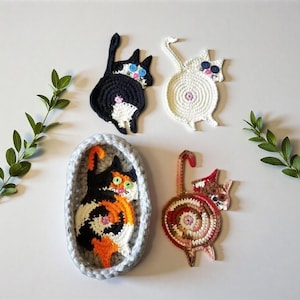 Könnte beinhalten: Gehäkelte Untersetzer in Katzenform in Schwarz, Weiß, Schildpatt und Rotbraun. Jede Katze hat Knopfaugen und ein einzigartiges Design. Eine Schildpattkatze befindet sich in einem grauen Korb. Dekorativ und funktional.