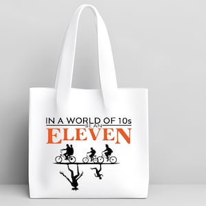 May include: White tote bag with black and orange text and graphics. The text reads "IN A WORLD OF 10s BE AN ELEVEN." Below the text are silhouettes of people riding bicycles and a mirrored image of the same. The bag has a white handle.