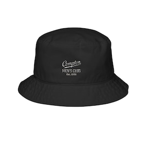 May include: Black bucket hat with a wide brim. The hat features the text "Compton MEN'S CLUB Est. 1983" in white script across the front. The hat is made of a smooth, solid black material and is suitable for casual wear.