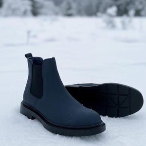 Op de afbeelding: Een paar marineblauwe Chelsea-laarzen met zwarte elastische zijpanelen en een dikke zwarte zool. De laarzen staan in een besneeuwde omgeving, wat hun ontwerp en constructie benadrukt.