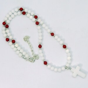 May include: A white beaded necklace with red accent beads and a white cross pendant. The necklace features a silver chain and clasp. The beads are round and evenly spaced, creating a simple yet elegant design.