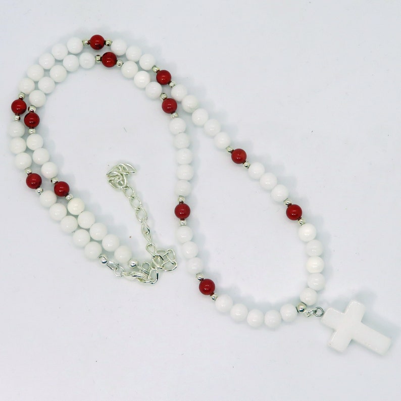 May include: A white beaded necklace with red accent beads and a white cross pendant. The necklace features a silver chain and clasp. The beads are round and evenly spaced, creating a simple yet elegant design.