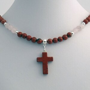 May include: A necklace featuring a cross pendant. The necklace has a strand of reddish-brown and pink beads, with silver accents. The cross pendant is a reddish-brown color and is suspended from the bead strand.