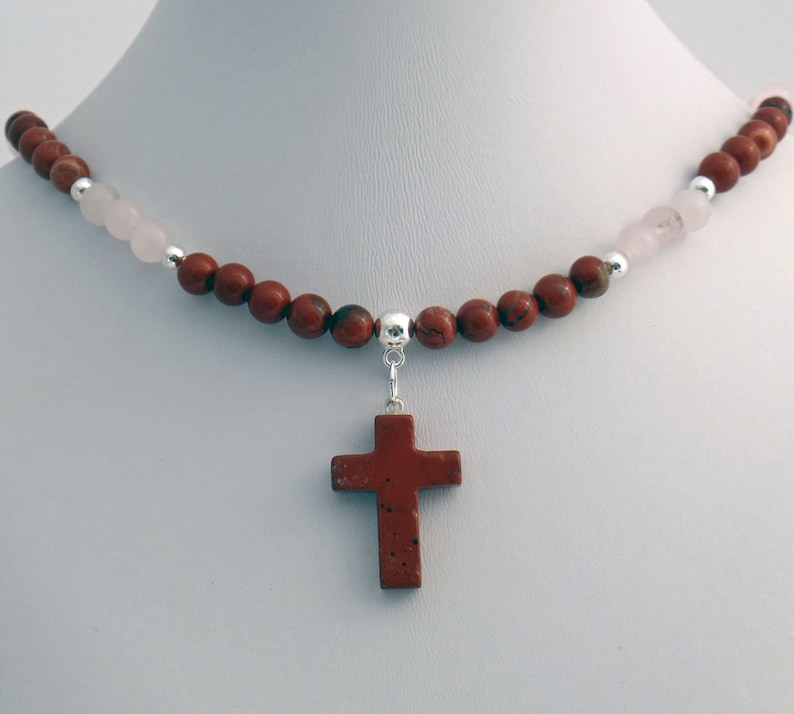 May include: A necklace featuring a cross pendant. The necklace has a strand of reddish-brown and pink beads, with silver accents. The cross pendant is a reddish-brown color and is suspended from the bead strand.