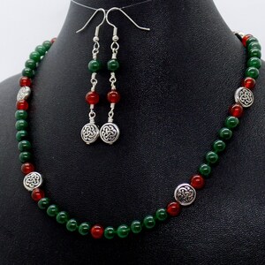 May include: A necklace and earring set featuring green and red beads. The necklace has silver-toned Celtic knot accents. The earrings have matching beads and Celtic knot charms. The jewelry is displayed on a black mannequin.