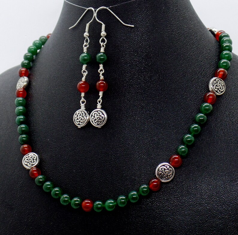 May include: A necklace and earring set featuring green and red beads. The necklace has silver-toned Celtic knot accents. The earrings have matching beads and Celtic knot charms. The jewelry is displayed on a black mannequin.